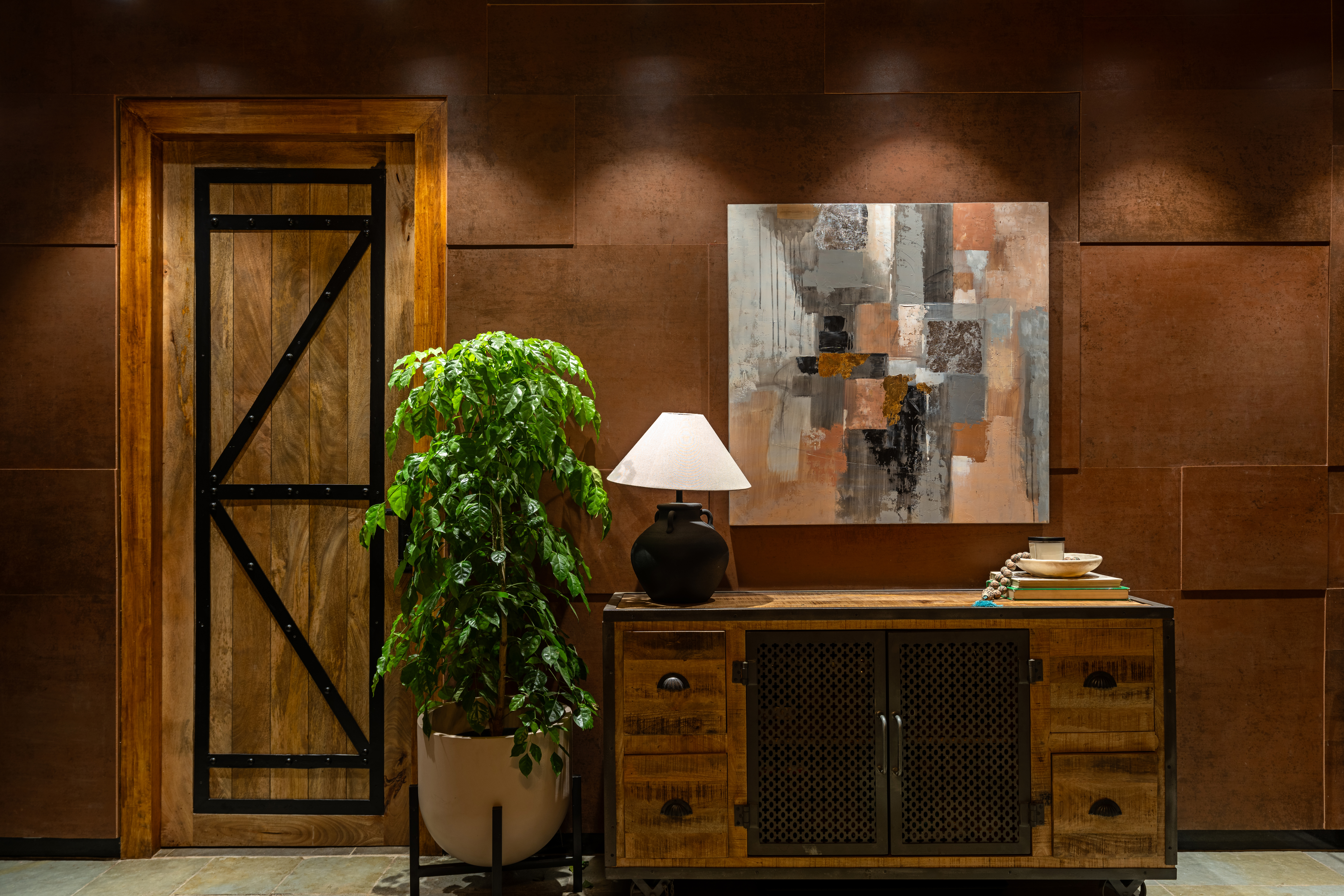 Timber console with mesh doors against a copper-panelled wall, holding a black urn lamp with a white shade, a stack of books and a bowl, beneath an abstract painting in the wall's own rust, copper and grey tones, with a plant in a white planter beside a plank door.